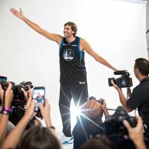 Dirk Nowitzki, in black jersey with Dallas Mavericks logo, poses for cameras amidst a frenzy of fans and flashbulbs. Stark white background