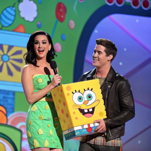 Katy Perry and Logan Henderson hosting the Nickelodeon Kids' Choice Awards. Katy holding a giant yellow spongebob. They're on a colorful, animated stage backdrop filled with cartoon elements.