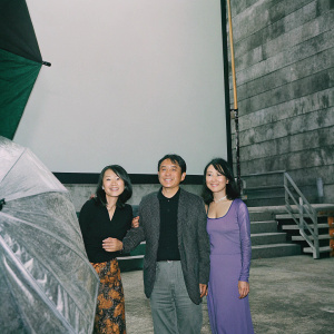 Jet Li, Jada, and Jane on the set of a Disney production. Jet Li in a black shirt and blazer, Jada in a vibrant dress, Jane in a purple dress. East Asian ethnicity. Warm smiles, against green screen backdrop.