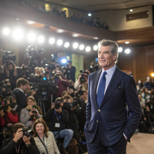 Pierce Brosnan, in a navy blazer and blue tie, at a film festival press conference. Crowded hall, many camera flashes.