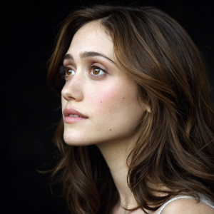 Medium close-up of Emmy Rossum, American actress. Brown shoulder-length hair, fair complexion, thoughtful expression. Dark blurred background emphasizes her features. Lighting highlights her face.