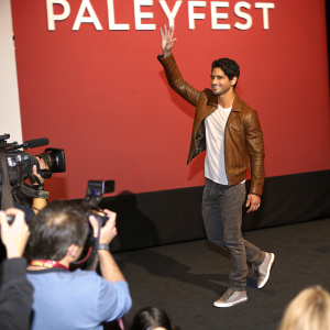 Tyler Posey waves at a bustling press conference. Wearing a brown leather jacket, gray sneakers, and a white t-shirt. The backdrop is a solid red wall with bold white text from Paleyfest.