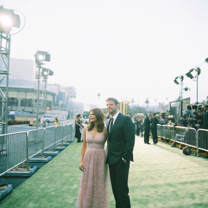 Danneel Ackles in a pastel pink, lace-trimmed dress, posing with Jensen Ackles in a black tuxedo at the Amazon Prime Awards. Flashing lights and a crowd of celebrities in the background.