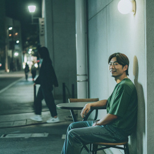 Kim Woobin, wearing a green t-shirt and jeans, at a casual cafe. Outdoor scene with street lamps and passerby