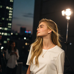 Brighton Sharbino, a young caucasian woman with long straight blonde hair, stands in a spotlight. She wears a simple white blouse and a subtle necklace. Against a blurred cityscape background, her serene expression is the focus of the candid photo.