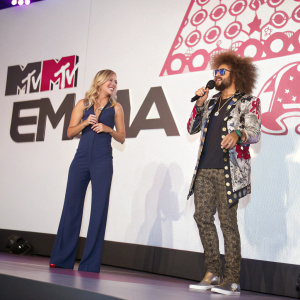 Laura Whitmore and Redfoo on stage at MTV EMA 2013. Laura in a sleek navy jumpsuit, Redfoo in a bohemian outfit. White backdrop, MTV and EMA logos visible.