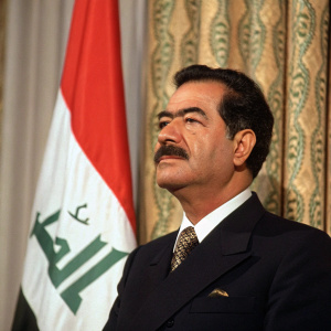 Saddam Hussein, with a mustache and wearing a dark suit, stands against a backdrop of a patterned curtain and Iraqi flag at a formal event.