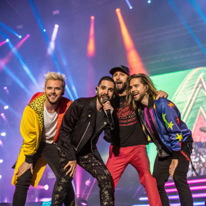 Members of Tokio Hotel, Gustav Schaefer, Bill Kaulitz, Tom Kaulitz, and Georg Listing, posing together on a vibrant stage setup at a concert.