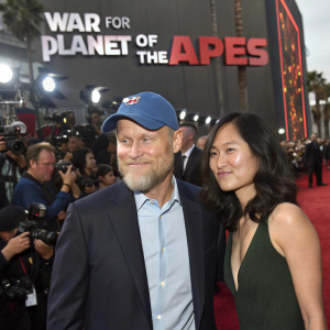 Caucasian actor Woody Harrelson, with a beard and blue baseball cap, alongside Asian actress Lara Louie, both wearing formal attire. They pose for photographers at the 'War for the Planet of the Apes' premiere event. Crowded red carpet scene, movie-themed backdrop visible.