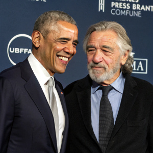 Barack Obama and Robert De Niro, both standing at a fundraising event. Obama has a warm smile, De Niro has a beard and a serious expression. Behind them is a dark blue background with corporate logos.