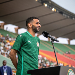 Riyad Mahrez, wearing a green Puma jersey, in a press conference. Stadium background with audience
