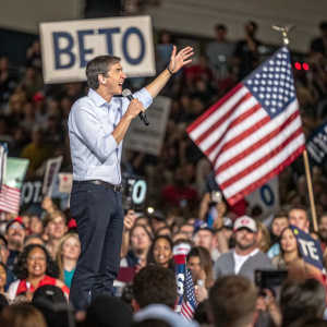Beto O'Rourke addressing an enthusiastic crowd at a campaign rally. The audience, diverse in ethnicity and gender, wave American flags and hold 'BETO' signs. Candid moment captured mid-speech.