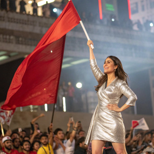Kareena Kapoor, Bollywood actress, in a shiny silver dress, waving a red flag. Crowd cheering in background