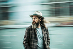 A middlenaged man with long hair and beard wearing a hat and a jacket, mysteriously