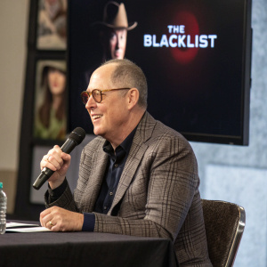 James Spader, actor, at a press conference. He sits confidently at a table, holding a microphone. Behind him is a large screen displaying the 'The Blacklist' logo