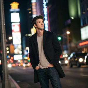 Grant Gustin stands confidently on a bustling street, wearing a black blazer over a casual t-shirt. Dark-washed jeans complete the look. Bright city lights and neon signs in the background