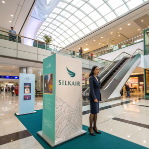 Promotional booth for SilkAir located in a busy mall. High ceilings with glass panels, escalators nearby. Stand features cut-out figure of a woman in professional attire. Informational panels with graphics and text about the airline's services.