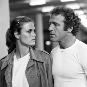 Barbara Trentham and James Caan from the film Rollerball. Black and white picture. James in a white t-shirt, Barbara in a brown jacket over a blouse. Styled ponytail and curly hair