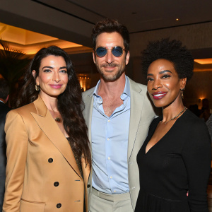 Shiva Rose McDermott, Dylan McDermott, and Lisa Gay Hamilton pose together at a glamorous party. They wear a tan blazer, light blue shirt with sunglasses, and black blouse respectively. Warm indoor lighting