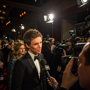 Eddie Redmayne, wearing a black suit and bow tie, giving an interview at a star-studded gala. Crowded, dimly lit room, multiple camera flashes