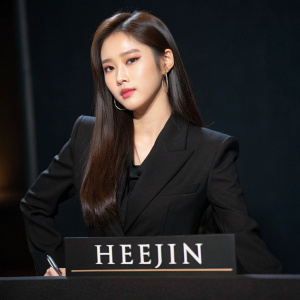 Heejin from Loona, Korean singer, confidently poses in a formal setting. Wearing a black blazer with a dark background. Her long straight brown hair and makeup are prominent. Signature below the photo
