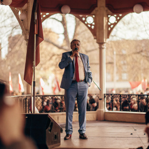 Jeremy Corbyn, in a blue suit with red tie, addressing a rally in a park. Labour Party banners flutter in the background.