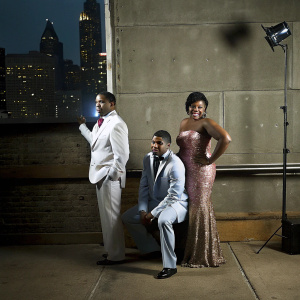 David Daniel Otunga Sr., David Daniel Otunga Jr., and Jennifer Hudson posing for a magazine cover shoot. Bright cityscape backdrop