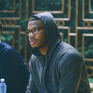 Damien Harris, running back for Alabama Crimson Tide, intense gaze. Gray hoodie, glasses. Dark backdrop with gold accents. Nearby water bottle.