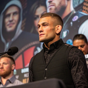 Dustin Poirier at an MMA conference. Focused look in a black vest. Fans and microphones in the background.
