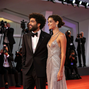 Noah Becker and Taina Moreno de Oliveira Lagoeiro at the Venice Film Festival. Becker in a black tuxedo, Lagoeiro in a silver gown. Dimly lit with red carpet and flashing cameras.