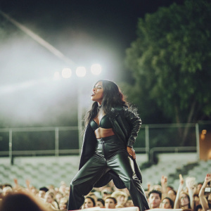 Sevyn Streeter on a packed concert stage, wearing a black leather outfit, performing energetically with a spotlight on her.