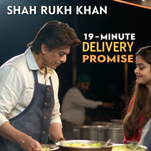Shah Rukh Khan in a bustling kitchen scene, alongside a woman with long hair. Promotional ad for grocery delivery service. Text in multiple languages highlighting 19 minute delivery promise. Dark background.