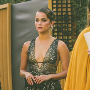 Close-up of Alicia Vikander at the Critics' Choice Awards. She is in a stunning black and gold dress. Serious expression. Gray backdrop with event details.