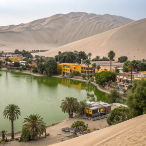 Serene photo of Huacachina village. Oasis-like setting with large, greenish lake. Colorful buildings in shades of yellow and other vibrant tones. Palm trees and vast sandy dunes in the background. Ideal for desert tourism