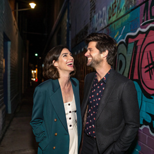 Lizzy Caplan and Adam Scott laughing at a party. Urban alleyway background with graffiti and dim lighting.