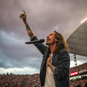 Brandon Boyd, lead singer of Incubus, is photographed mid-performance at a packed stadium concert. His tattoos are visible as he holds the microphone with one hand, the other raised to the crowd.