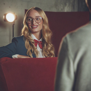 Sophie Turner, long blonde hair, friendly smile, wearing a navy blue blazer and a white blouse with a red bow tie, posing for a semi-formal event photo in a studio with plain backdrop