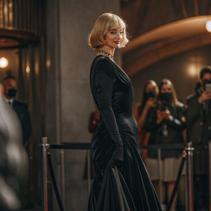 Krysten Ritter, fair-skinned, short blonde bob, standing confidently. Wearing a black velvet dress. Golden globe awards red carpet, blurred crowd in background.