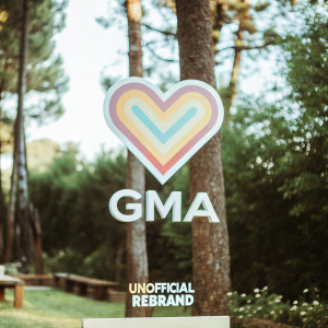 An unofficial redesign of the GMA logo. The new look features a vibrant heart symbol in a spectrum of colors against a deep purple backdrop. Below the heart, the GMA name is displayed in a sleek, modern font. A smaller note at the bottom reads 'Unofficial Rebrand'