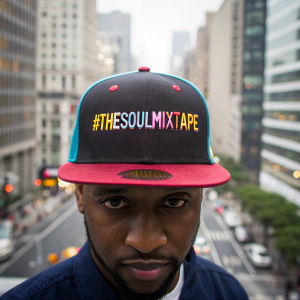 Close up of trendy Black man's head, facing camera, with a cityscape background. Hes wearing a trendy baseball cap with the message "#TheSoulMixtape" on the front of the cap in colourful 1970's font