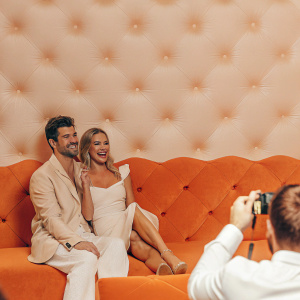 Kristin Cavallari and Nick Zano at a glamorous awards show. They are seated together on a velvet sofa, beaming with smiles. The backdrop is a sparkling diamond pattern, with a sea of photographers in the foreground.