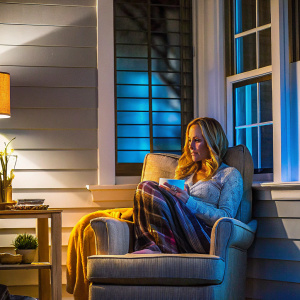 Kim Raver from Grey's Anatomy photographed in a cozy, minimalist living room. Soft natural light illuminates her long blonde hair. She wears a relaxed outfit and sits on a plush armchair.