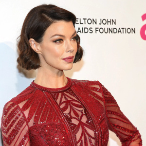 Close-up portrait of actress Pollyanna McIntosh at Elton John AIDS Foundation gala. She's wearing a red sequined dress with intricate patterns. White backdrop, pink logo, celebration of an anniversary