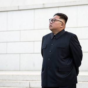Kim Jong Un, Asian leader, wearing a dark suit and round glasses. Solemn pose, standing in front of a plain white wall.