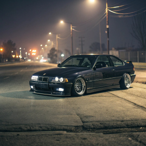 Custom BMW E36 M3, sleek midnight-black with chrome accents, parked on a deserted street at night. Custom suspension highlights the car's sporty and powerful demeanor.