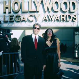 Ron Perlman and Opal Perlman attending a press conference. Ron in a black suit with a white shirt and red tie, Opal in a stylish black dress. Event backdrop features the title 'Hollywood Legacy Awards'