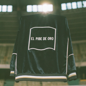 Diego Maradona's iconic number 10 jersey on display. The black jersey with white accents featuring Maradona's 'El Pibe de Oro' logo is framed against a blurred stadium backdrop.