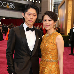 Takamasa Ishihara and Melody Miyuki Ishikawa attending the Academy Awards. Takamasa in a sleek tuxedo, Melody in an ornate gold dress. Glamorous red carpet setting.
