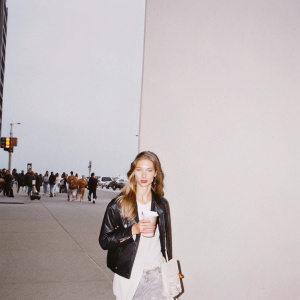 Rosie Huntington-Whiteley on a busy city street. Wearing casual attire, a black leather jacket over a white shirt. Holding coffee. Background is morning rush hour crowd.