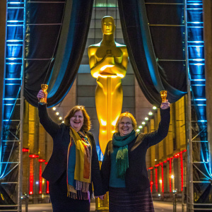 Fiona Crombie and Finola Dwyer celebrating their Oscar win. They stand in front of a golden statue. Large curtain backdrop